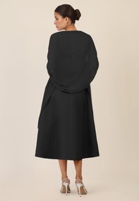 Vestido largo negro con tela fluida, con un escote barco y una cintura ajustada. La modelo lleva tacones plateados, que muestran una artesanía sencilla.