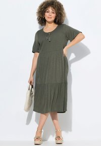 Vestido maxi verde oliva con mangas cortas, cuerpo texturizado, detalle atado en el escote y falda acampanada. Combinado con un bolso de tejido y sandalias de plataforma.