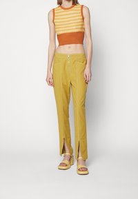 Stickad crop-top med gula och orange randningar, kombinerad med senapsgula byxor med fram-slitsar och dragkedjedetaljer. Gula klackskor med blockklack.