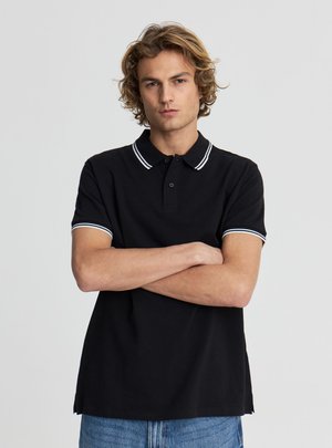 Młody mężczyzna o kręconych włosach, ubrany w czarną koszulkę polo z białym wykończeniem, z założonymi ramionami, stojący na jednolitym jasnoszarym tle.