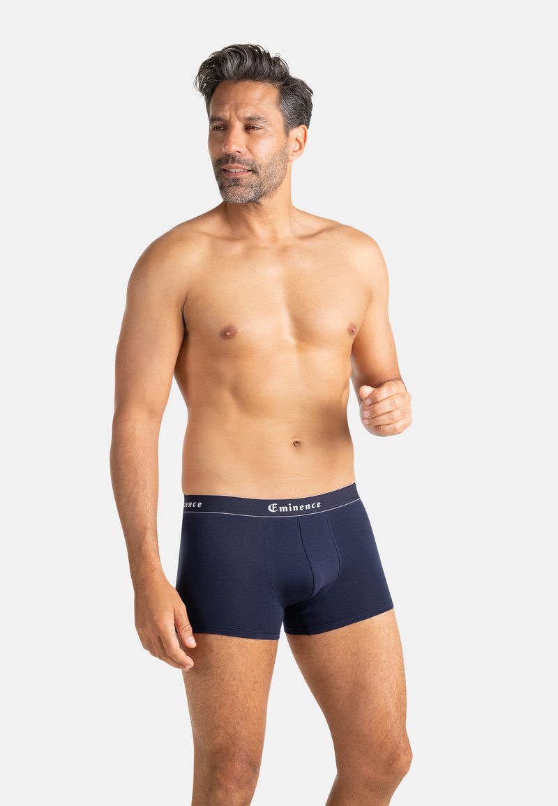 Homme torse nu aux cheveux courts et foncés portant un boxer Eminence bleu marine, debout devant un fond clair uni.