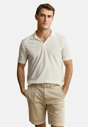 Jonge man die een wit poloshirt met korte mouwen en beige korte broek met riem draagt, staand met handen in de zakken tegen een effen achtergrond.