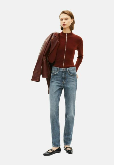 Top zip con lavorazione a maglia color bordeaux, jeans blu a gamba dritta, ballerine nere con dettaglio a fiocco e una giacca di pelle marrone appoggiata sulla spalla.