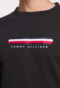 Sudadera negra con cuello redondo, que presenta una franja de logotipo horizontal en rojo, blanco y azul, junto con el texto "TOMMY HILFIGER" en blanco.