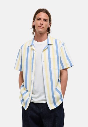 Homme aux cheveux mi-longs portant une chemise à manches courtes à rayures verticales bleues, jaunes et blanches par-dessus un t-shirt blanc.