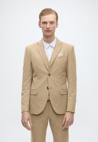 Costum bej deschis din țesătură texturată, cu rever tăiat, două butoane și un batist alb în buzunar. Asortat cu o cămașă albă.