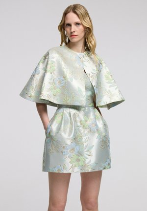 Blonde vrouw die een pastel bloemrijke brokaat capelet draagt over een bijpassende korte jurk met zakken, staand tegen een effen achtergrond.