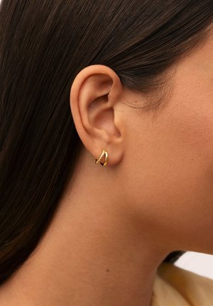 Orecchio destro di una donna con capelli scuri riposti dietro, che indossa un piccolo orecchino a cerchio dorato attorcigliato su uno sfondo neutro.