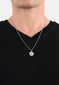 Collana in argento con ciondolo rotondo che presenta un design di cervo e un piccolo accento. Indossata sopra una t-shirt nera a scollo a V.