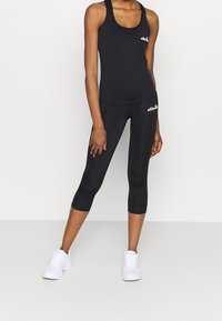 Donna che indossa un top senza maniche nero di ellesse e leggings corti con scarpe da ginnastica bianche, in piedi di fronte a uno sfondo chiaro e uniforme.
