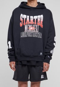 Sudadera negra con estampado gráfico en rojo y blanco que dice "STARTER MCMXCI" y "LOOKFORTHESTAR". Cuenta con un bolsillo frontal y puños acanalados. Se combina con pantalones cortos de malla negros.