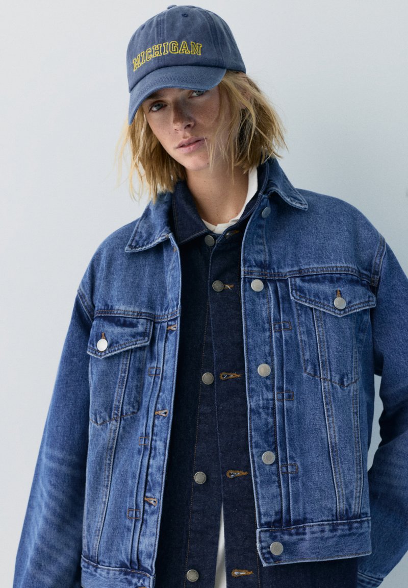 Personne portant des vestes en jean bleu superposées et une casquette bleu marine avec des lettres jaunes "Michigan" sur un fond uni.