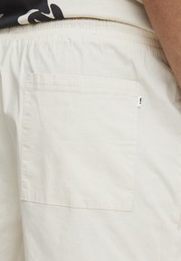 Pantalones cortos de color beige claro, hechos de algodón, con una cintura fruncida y un bolsillo trasero rectangular. Textura suave, diseño minimalista.