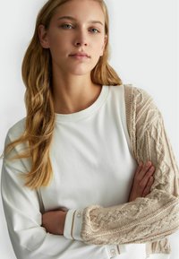 Felpa caratterizzata da un corpo bianco, maniche in maglia beige testurizzata con motivi a treccia, collo rotondo e polsini a costine con dettagli del logo.