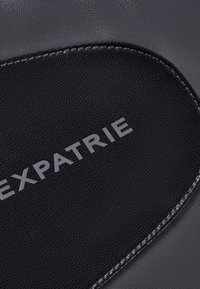 Čierna kožená taška s textúrovaným povrchom, so šedým prešívaním a slovom "EXPATRIE" v modernom písme. Mierne zakrivený dizajnový detail.