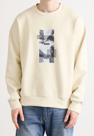 Beżowa bluza z grafiką czarno-białej panoramy miasta i napisem "BOSS" na środku, noszona przez stojącą osobę w niebieskich dżinsach.