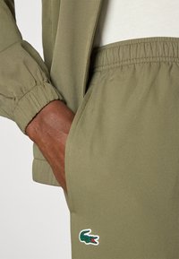 Ελαιόχρωμο αθλητικό κοστούμι με ελαστικές μανσέτες, λεία υφή και λογότυπο κροκόδειλου στη τσέπη. Κοντινό πλάνο του χεριού που κρατάει τη λεπτομέρεια της τσέπης.