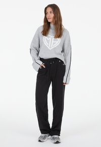 Hellgrauer gestrickter Pullover mit einem Herzdesign, kombiniert mit schwarzen Jogginghosen mit Seitentaschen und einem Kordelzugbund. Graue Sneakers.