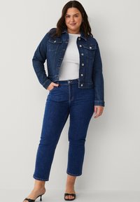 Chaqueta de mezclilla en azul oscuro con cierre de botones y dos bolsillos en el pecho, combinada con jeans ajustados azul oscuro y sandalias de tacón negro.