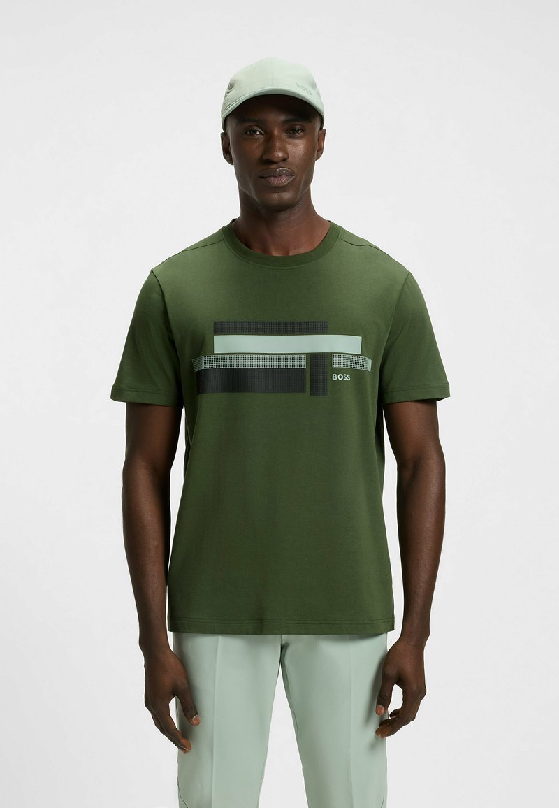 Grünes Baumwoll-T-Shirt mit einem geometrischen Grafikdesign und schwarzen sowie hellgrünen Streifen. Mit Rundhalsausschnitt und kurzen Ärmeln gestaltet.