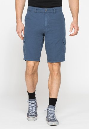 Uomo con pantaloncini cargo blu, calzini neri e scarpe da ginnastica alte grigie, in piedi su uno sfondo bianco.