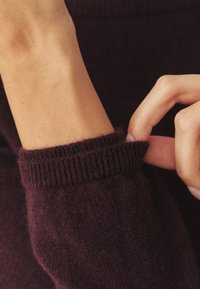 Burgundijas adīta džempera piedurkne ar ribotu apmali. Tekstūra izskatās maiga un pūkaina, ar bezšuvju dizainu ap plaukstas daļu.