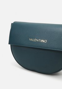 Tyrkysová kožená kabelka s kulatou klopou, zlatým kováním s logem „VALENTINO“ a hladkým povrchem.