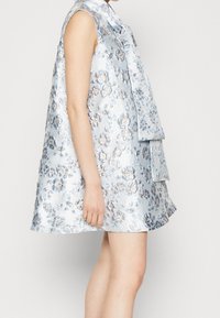 Sininen kukkakuosiset hihaton mekko kiiltävällä satiinipinnoitteella, jossa on kerrokselliset röyhelöt ja löyhä A-linjainen muoto.