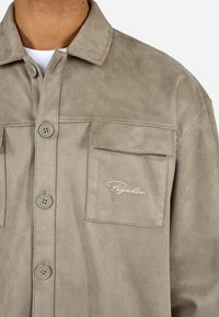 Beige műsuede dzseki gombos záródással, két mellzsebbel és hímzett logóval a jobb zsebben. Puha tapintás és laza dizájn.