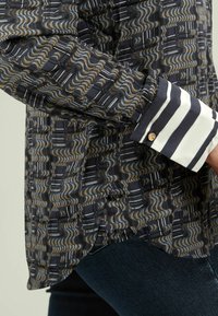 Camicia a maniche lunghe con un tessuto scuro a motivo ondulato nei toni del blu, oro e bianco. Presenta un polsino a strisce bianco e navy con un dettaglio in oro.