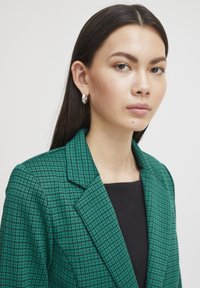 Zelený kostkovaný blazer s klopovým límcem, vyrobený z texturované tkaniny. Nosený přes černý top, doplněný stříbrnými kruhovými náušnicemi pro zvýraznění.