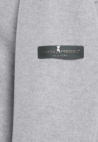 Lichtgrijs vilt met een donkergroene rechthoekige label met de tekst "FRIEDA FREDDIES® NEW YORK" en een klein logo van een hond.