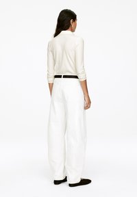 Woman wearing white ribbed long-sleeve shirt, wide white pants with black belt, and black shoes, standing facing away on white background.