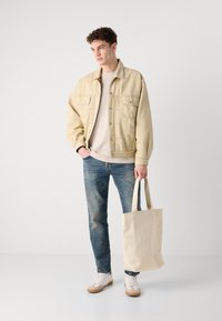 Jachetă din denim bej peste un pulover deschis, asortată cu blugi albaștri. Ținând o geantă crem, purtând adidași albi cu tălpi bej.