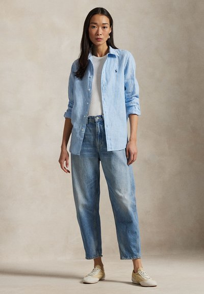 Tânără în picioare, purtând o cămașă albastră cu dungi peste un tricou alb, blugi albaștri deschiși cu talie înaltă și pantofi sport bej, pe un fundal simplu.