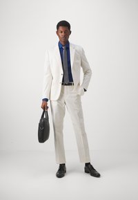 Abito chiaro con camicia blu e cravatta grigia. Scarpe in pelle nere e una borsa nera textured. Slim fit con design su misura.