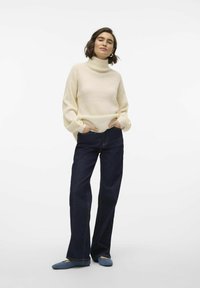 Krämfärgad polokrage tröja med ribbad textur, i kombination med vida mörk denimjeans och blårandiga platta skor, händerna i fickorna, neutral bakgrund.