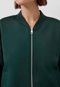Grüne Bomberjacke mit glatter Textur, geripptem Kragen, Reißverschluss und seitlichen Taschen. Besitzt ein minimalistisches Design ohne Muster.