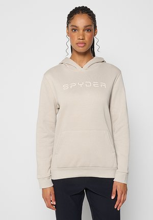 Frau mit geflochtenen Haaren, die einen beigen Spyder-Hoodie und schwarze Hosen trägt, steht vor einem schlichten hellen Hintergrund.