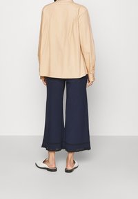 Personne portant un haut beige à manches longues, un pantalon large en ribbed bleu marine avec une bordure en dentelle, et des chaussures blanches sans lacets, se tenant de dos sur un fond uni.