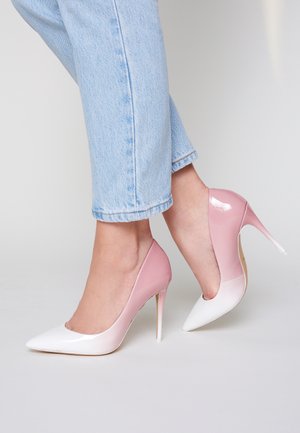 Feet wearing light blue jeans and two-tone pink and white high-heeled stilettos, standing on a light gray surface.