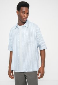 Ανοιχτό μπλε κοντομάνικο πουκάμισο με κάθετες ρίγες, τσέπη στο στήθος και άνετη εφαρμογή, φορεμένο πάνω από ένα ανοιχτό μπλε εσώρουχο.