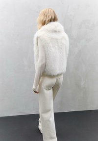 Weißer Kunstfell-Westen mit hohem Kragen, getragen über einem leichten, taillierten Oberteil mit langen Ärmeln, kombiniert mit hellen, weit geschnittenen Hosen.