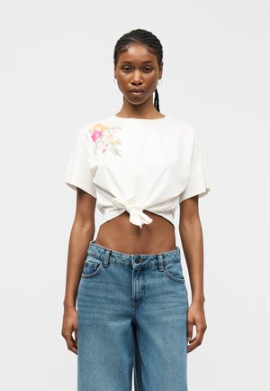 Mujer con top corto de manga corta blanco con bordado floral y lazo en la parte delantera, combinado con vaqueros azules de tiro alto.