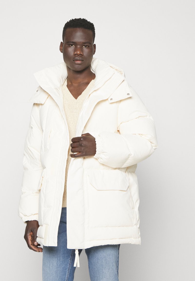 Doudoune en tissu crème avec capuche, dotée de poignets élastiques, de poches avant et d'une fermeture éclair. Stylée par-dessus un pull beige et un jean.