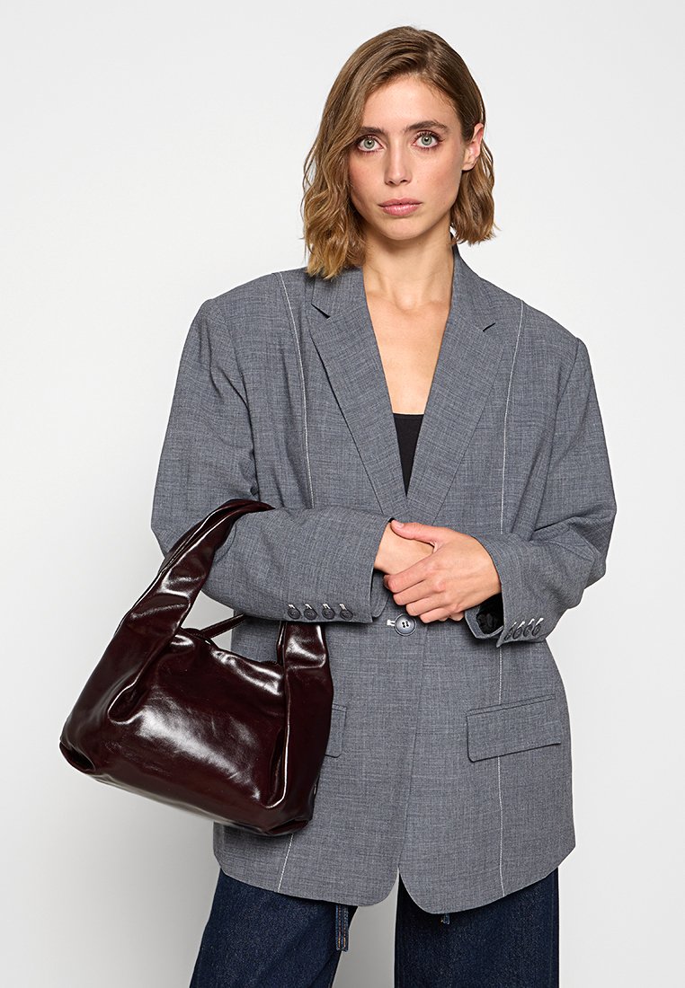 Blazer gris avec des coutures visibles, accompagné d'un sac à main en cuir verni bordeaux. Modèle portant un haut noir et un jean en denim.