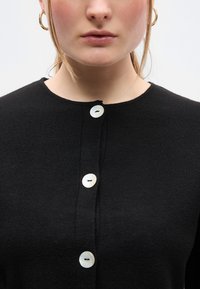 Μαύρη πλεκτή ζακέτα με στρογγυλή λαιμόκοψη, τέσσερα λευκά κουμπιά και υφή στο ύφασμα. Τα κουμπιά έχουν ελαφριά γυαλάδα και ραφτά διακοσμητικά λεπτομερή.