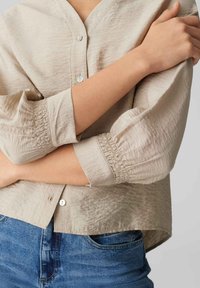 Beige button-up shirt with a textured fabric, featuring elasticized cuffs and a relaxed fit. Paired with blue jeans.