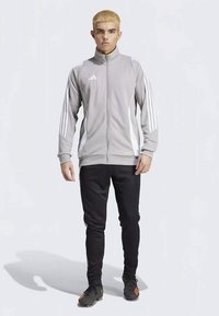 Grau sportliche Jacke mit weißen seitlichen Streifen und einem Reißverschluss vorne, kombiniert mit schwarzen schmal geschnittenen Hosen und schwarzen Schuhen mit orangenen Akzenten.