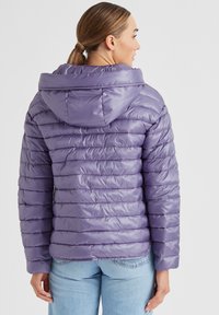 Lila gepolsterte Jacke mit Kapuze, mit horizontaler Steppung, glänzender Textur und elastischen Bündchen. Getragen mit hellblauen Jeans.